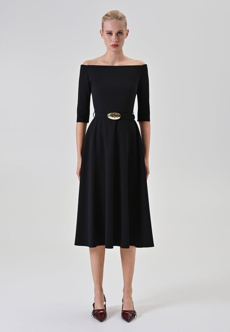 Schwarzes schulterfreies Kleid mit drei Viertelärmeln, tailliertem Oberteil, fließendem Rock und einer goldenen Gürtelrose. Hat eine glatte Textur.