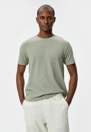 Camiseta verde de manga corta con tejido texturizado, escote redondo y corte holgado, combinada con pantalones de color claro.