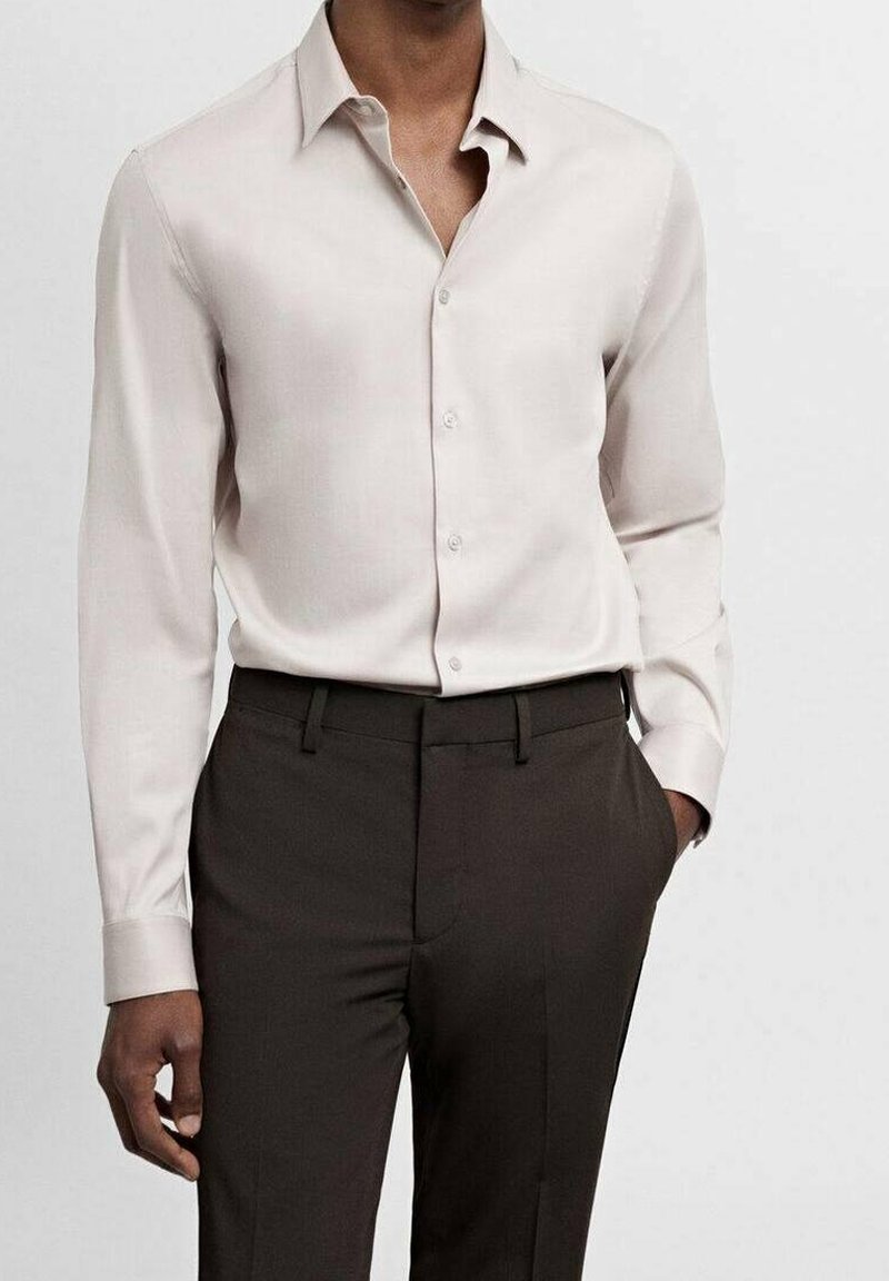 Homme portant une chemise beige à manches longues boutonnée, rentrée dans un pantalon habillé marron foncé, une main dans la poche, sur fond blanc.