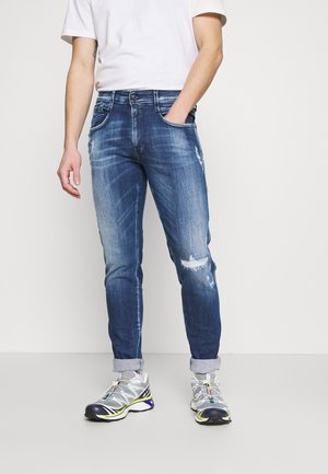 Hombre con vaqueros azules desgastados remangados con parches desgastados y zapatos deportivos grises contra un fondo blanco liso.