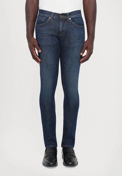 Jeans in denim blu scuro con vestibilità slim, cinque tasche e cuciture chiare, abbinati a scarpe nere lucide.