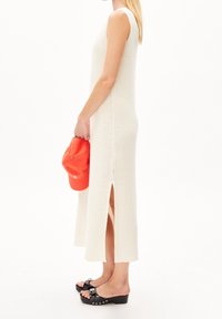 Robe tricotée, sans manches, de couleur crème avec des fentes sur les côtés ; associée à une casquette orange vif et des sandales à plateforme noires ornées de clous décoratifs.