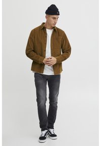 Smeđi kord č shirt s bijelom majicom ispod, uparen s tamnim trapericama i crnim tenisicama. Crna kapa za uši upotpunjuje izgled.
