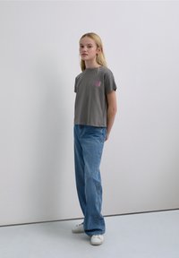 Gray short-sleeve t-shirt with a pink graphic, paired with light blue jeans featuring star-shaped embroidery. Model stands against a plain backdrop.