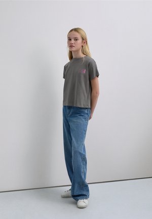 Gray short-sleeve t-shirt with a pink graphic, paired with light blue jeans featuring star-shaped embroidery. Model stands against a plain backdrop.