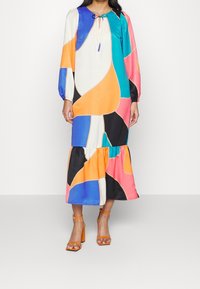 Femme portant une longue robe à motif abstrait multicolore à manches longues, assortie à des sandales à talons blocs orange.