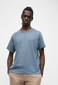 Νεαρός άνδρας με κοντά dreadlocks στέκεται με τα χέρια στις τσέπες, φορώντας μπλε μπλούζα και μπεζ παντελόνι, σε απλό λευκό φόντο.