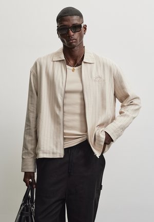 Mand iført stribet beige jakke over tanfarvet skjorte, sorte bukser, solbriller, halskæde, der holder sort taske med den ene hånd i lommen.