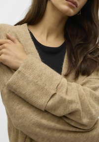 Beige Strickcardigan mit gerippten Bündchen, tiefem V-Ausschnitt und weicher Textur, über einem schwarzen, gerippten Oberteil getragen.