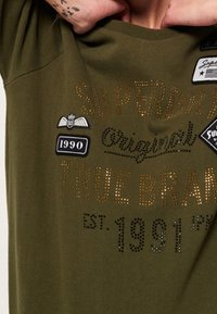 Camiseta verde oliva con texto metálico dorado y parches negros. El diseño incluye "SUPERDRY", "1990" y "EST. 1991" en varias fuentes.