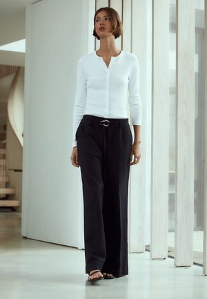 Femeie în picioare în interior, purtând un top alb cu mâneci lungi cu nasturi, pantaloni negri cu croială largă și curea, și sandale negre.