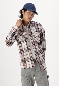Flanellen shirt met een oranje, marineblauwe en crèmekleurige plaidpatroon; heeft twee borstzakken en een knoopsluiting. Gecombineerd met een grijze cargobroek en een marineblauwe pet.