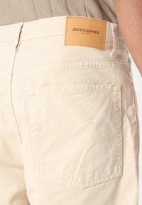 Pantalones cortos de algodón beige con una etiqueta de cuero marrón que dice "JACK & JONES". Presenta un bolsillo trasero con un detalle de costura curva.