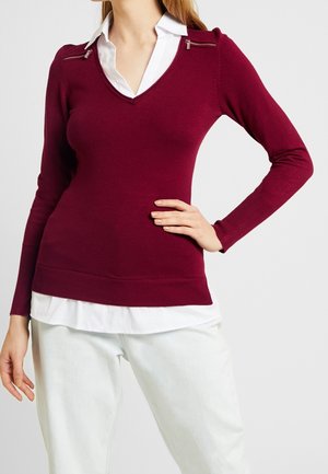 Bordeaux v-hals trui met lange mouwen, voorzien van zilveren ritsaccenten op de schouders en een geribbelde zoom. Gelaagd over een wit shirt.