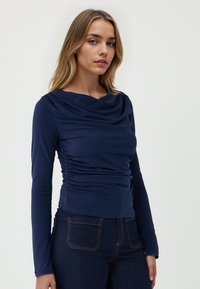 Maglia a maniche lunghe blu navy con scollatura drappeggiata e dettaglio arricciato di lato, abbinata a jeans in denim scuro con cuciture dorate.
