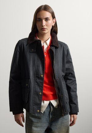 Junge Frau trägt eine schwarze Jacke mit dunklem Kragen, roten Pullover, weißes Hemd mit Kragen und blaue Jeans vor einem einfarbigen hellen Hintergrund.