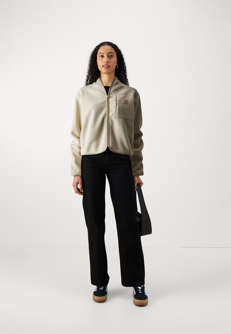 Beige fleece jack met een staande kraag, voorrits en borstzak, gecombineerd met zwarte wijde broek en donkere sneakers.