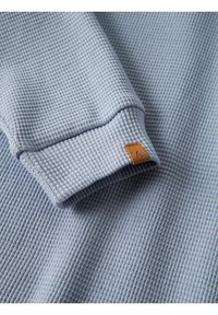 Tissu en maille gaufrée bleu clair avec un revers de manche plié présentant une petite étiquette en cuir marron cousue sur le bord.