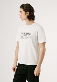 Balta kokvilnas T-kreklu ar tumši zilu grafisku printu "PEPE JEANS LONDON" priekšā, īsām piedurknēm un apaļu kakla izgriezumu. Apvienots ar melniem biksēm.