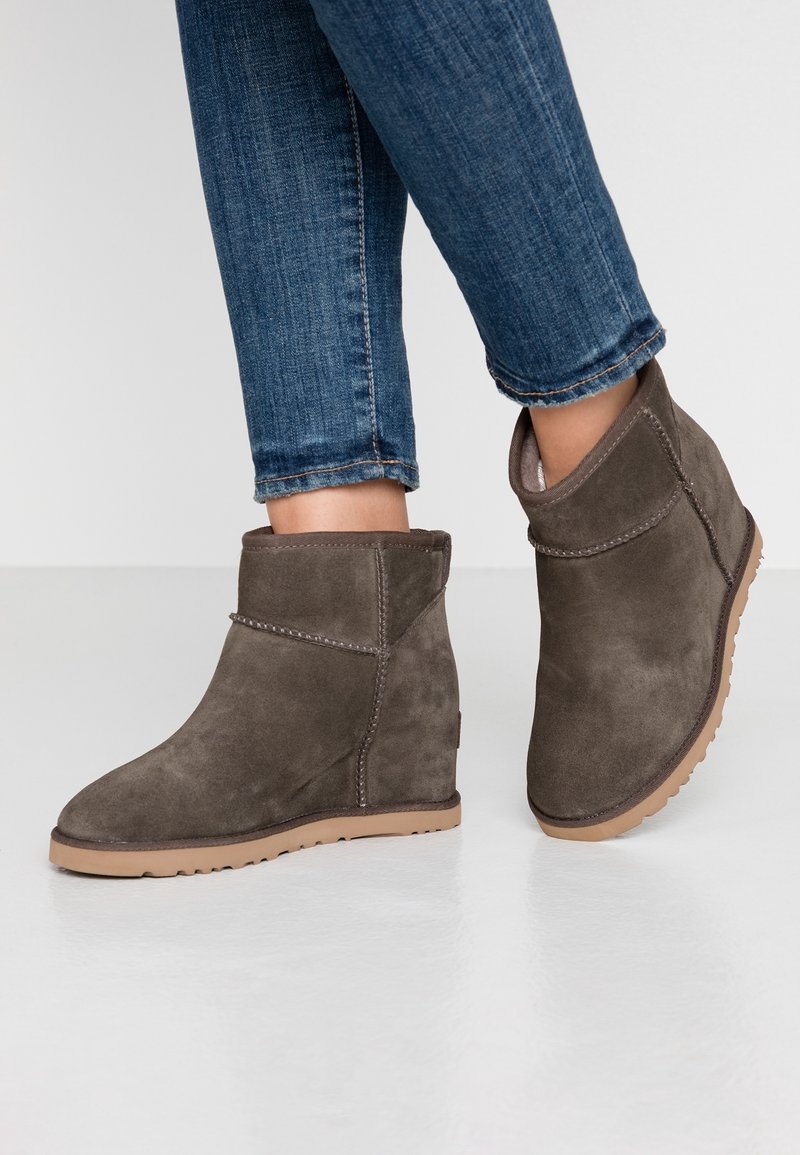 Botas de tobillo de ante marrón con cuña, con detalles de costura y puntera redondeada, combinadas con jeans ajustados azules, mostrando un look casual.