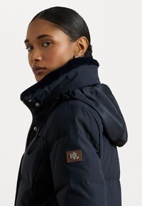 Tamsiai mėlynas pūkinis švarkas su dirbtiniu kailiniu gaubtu, užtrauktuku ir spaudėmis, o kairiajame rankovėje yra rudos odos emblema su inicialais.