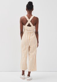Combinaison rayée avec des rayures verticales orange et blanches. Présente des bretelles croisées dans le dos, un détail découpé et des jambes larges raccourcies. Tissu doux.