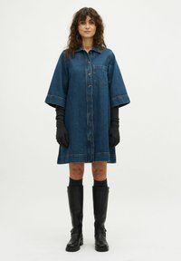 Dunkelblauses Denim Kleid Hemd mit kurzen Ärmeln, Knopfverschluss, Brusttasche und lockerer Passform. Kombiniert mit schwarzen knielangen Stiefeln.