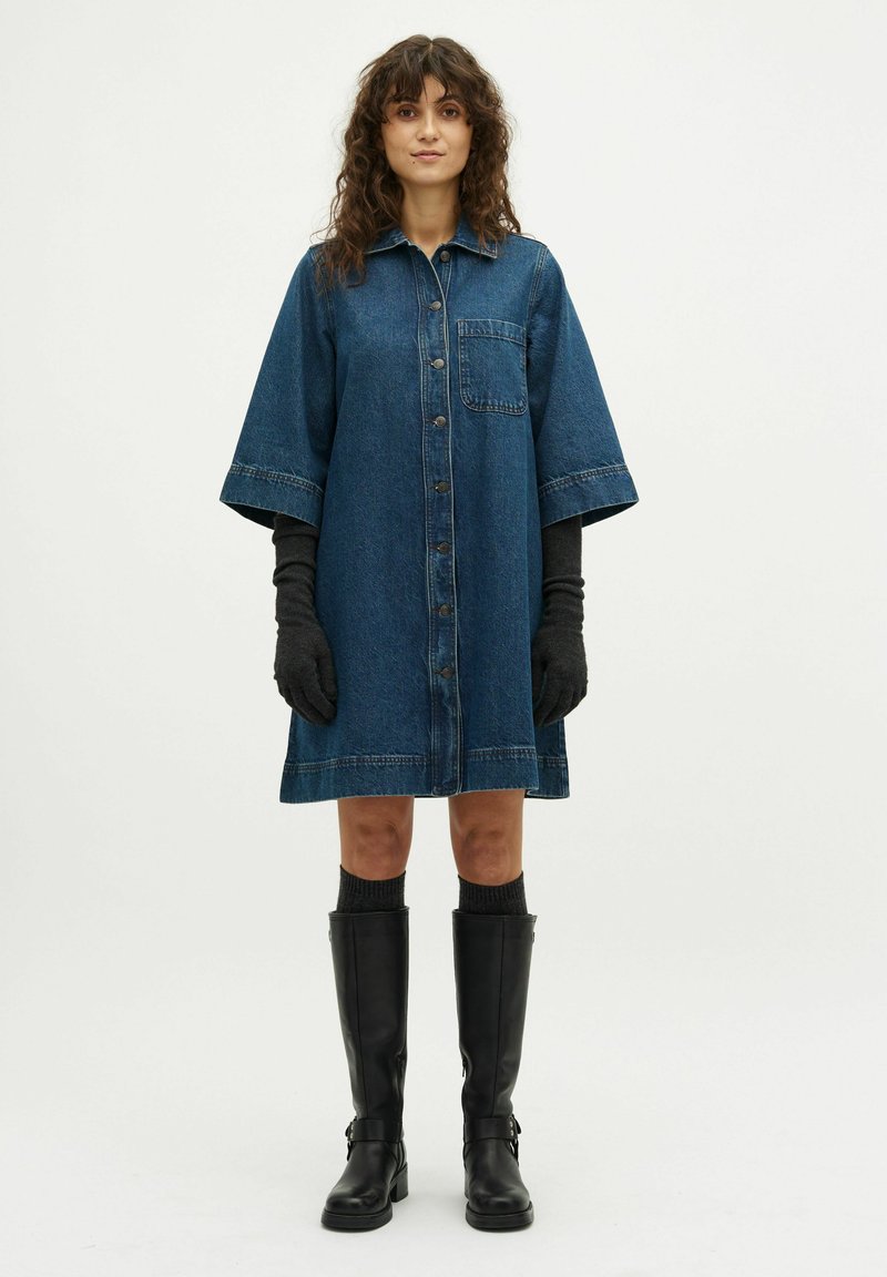 Dunkelblauses Denim Kleid Hemd mit kurzen Ärmeln, Knopfverschluss, Brusttasche und lockerer Passform. Kombiniert mit schwarzen knielangen Stiefeln.