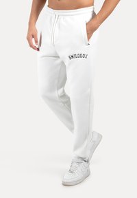 Bílé bavlněné tepláky s elastickým pasem, šňůrkou na stažení, bočními kapsami a černým výšivkou "SMILODOX" na horní levé stehně.