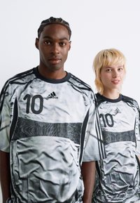 Dos personas vistiendo camisetas de fútbol Adidas a juego con el número 10, de pie frente a un fondo blanco liso.