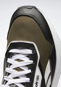 Černo-olivová teniska s horní částí z texturované tkaniny, bílými tkaničkami a bílým logem na boku. Zakřivená podrážka a polstrovaný límec.