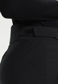 Black high-waisted pants with a wide waistband featuring Velcro closure, smooth texture, and subtle seam detailing on the sides.