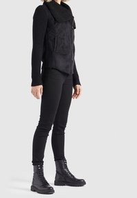 Personne portant une veste noire à fermeture éclair avec un grand col, un pantalon noir ajusté et des bottes de combat noires à lacets, debout devant un fond uni.