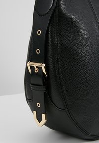 Sac en cuir texturé noir avec une sangle noire réglable dotée de boucles en métal doré et de détails matériels sur un fond neutre.