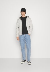 Light gray zip-up jacket with black drawstrings, black t-shirt, light blue jeans, and black and white sneakers, standing against a white background.