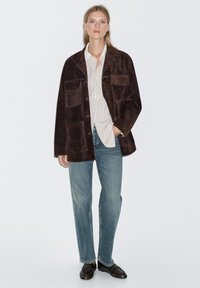 Braune Wildlederjacke mit zwei Brusttaschen, kombiniert mit einem weißen, gestreiften Hemd, hellblauen Jeans und braunen Schuhen. Einfaches Design, lockere Passform.