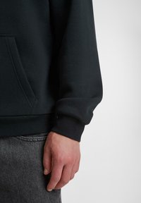Schwarzer Sweatshirt mit lockerer Passform, gerippten Bündchen und Kängurutasche. Glatte Textur mit subtiler glänzender Oberfläche. Hand ruht auf dunklen Jeans.