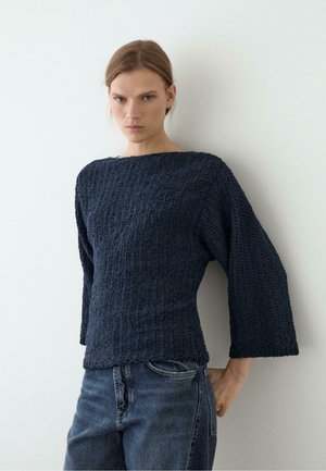 Frau mit hellbraunen Haaren trägt einen marineblauen texturierten Pullover mit weiten Ärmeln und blaue Jeans, steht vor einer weißen Wand.