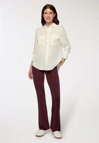 Blusa bianca a maniche lunghe con due tasche sul petto, abbinata a pantaloni a campana color bordeaux e sneakers bianche, su sfondo neutro.