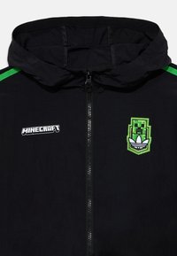 Čierna kapucňová bunda s zelenými detailmi a brandingom Minecraft, so zipsom vpredu a textúrovanou látkou. Vyšité logo na hrudi.