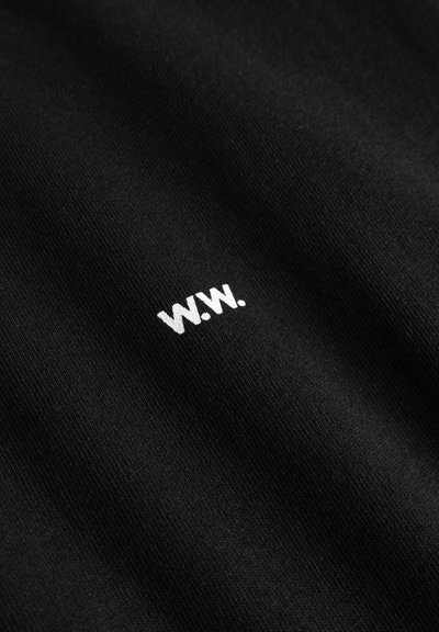 Tecido preto com uma textura suave apresenta um pequeno logo bordado branco "W.W." posicionado de forma subtil. O design é minimalista e contemporâneo.