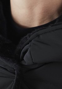 Chaqueta acolchada negra con un suave cuello de piel sintética. Presenta un cierre de cremallera y un diseño recogido cerca del cuello para mayor calidez.