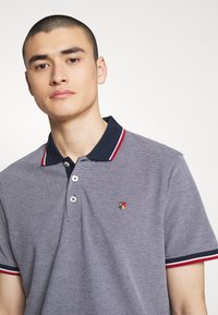 Polo majica v mornarsko modri in beli barvi z rdečimi in modrimi črtastimi ovratniki. Ima gumbe in majhen večbarven logotip na prsih. Teksturirana tkanina.