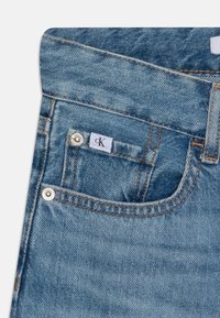 Jasnoniebieskie dżinsy z małą przednią kieszenią z widoczną metką, wzmocnionymi szwami i metalowymi nitami. Miękka, teksturowana powierzchnia.