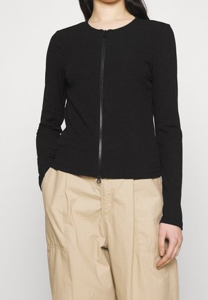 Frau trägt ein eng anliegendes, schwarzes, langärmeliges Zip-up-Oberteil und lockere beige Hosen, steht mit den Händen in den Taschen vor weißem Hintergrund.