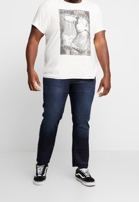 White t-shirt with a gray graphic print, dark blue denim jeans, and black sneakers with white accents. Casual fit and short sleeves.
