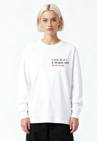 Donna bionda con capelli corti che indossa una maglietta bianca a maniche lunghe con la scritta "Un po' di [cuore] in questo mondo brutale (ma non troppo)"