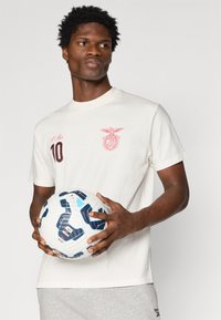 Λευκό μπλουζάκι από βαμβάκι με κόκκινο έμβλημα και τον αριθμό 10, συνοδευόμενο από γκρίζες φόρμες. Το μοντέλο κρατάει μια λευκή μπάλα ποδοσφαίρου με μπλε και μαύρες λεπτομέρειες.