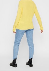 Person med en gul stickad tröja, ljusblå jeans och svarta ankelstövlar, står med ryggen vänd mot betraktaren.