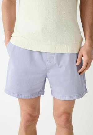 Mann trägt hellviolette Shorts mit Taschen und ein cremefarbenes kurzärmeliges Strickshirt, steht vor einem weißen Hintergrund.